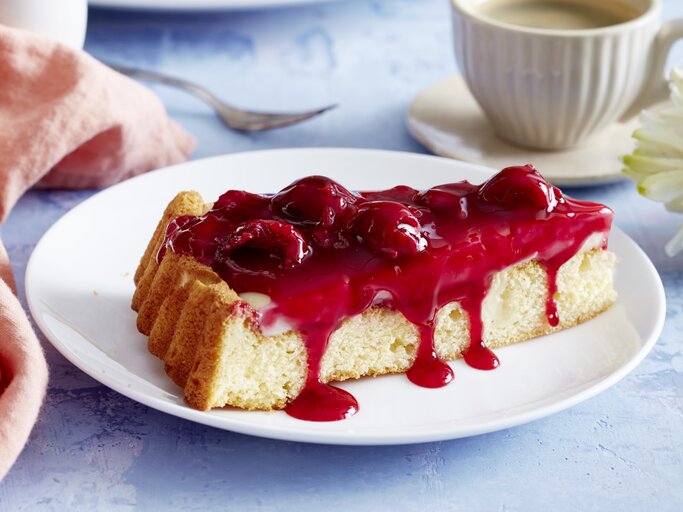 Pudding-Kirschkuchen auf einem weißen Teller, blauer Untergrund und Geschirr und Küchentuch im Hintergrund.  | © EAT CLUB / Isabell Triemer