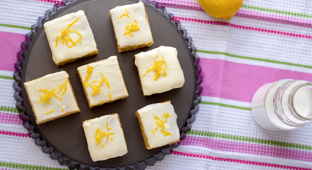 Mehrere Stücke von den Zitronenschnitten mit Limoncello-Guss auf einem dunklen Teller. Darunter eine bunte Tischdecke mit grünen und pinken Streifen.