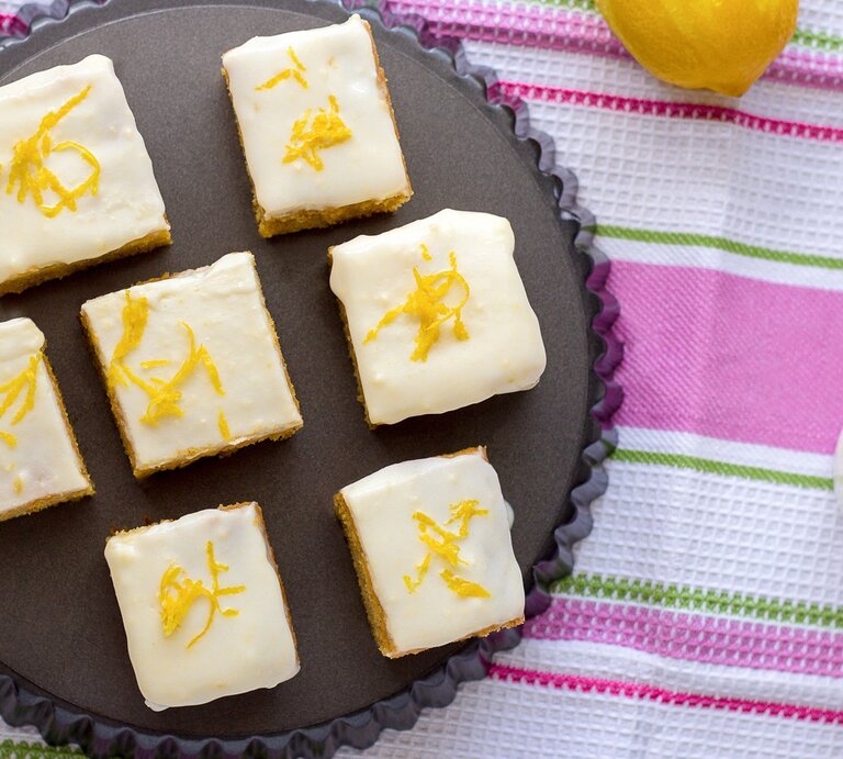 Mehrere Stücke von den Zitronenschnitten mit Limoncello-Guss auf einem dunklen Teller. Darunter eine bunte Tischdecke mit grünen und pinken Streifen. | © Shutterstock/ Juliya_Ka