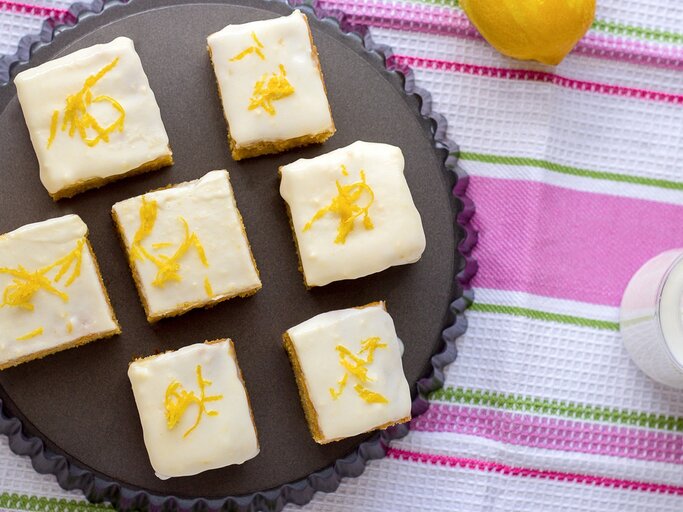 Mehrere Stücke von den Zitronenschnitten mit Limoncello-Guss auf einem dunklen Teller. Darunter eine bunte Tischdecke mit grünen und pinken Streifen. | © Shutterstock/ Juliya_Ka