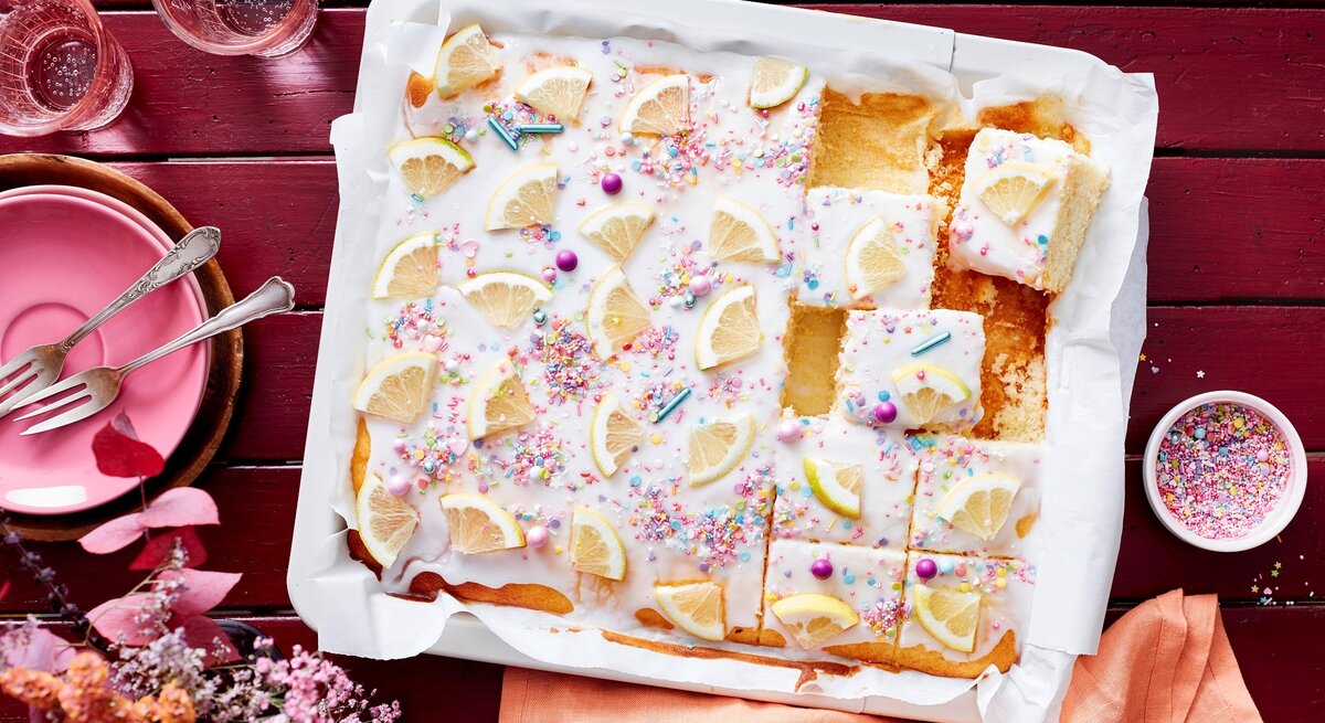 Auf einem weißen Teller liegt ein saftiger Zitronen-Buttermilchkuchen. Er ist mit Puderzuckerglasur und Zuckerstreuseln dekoriert und mit frischen Zitronen bedeckt. 