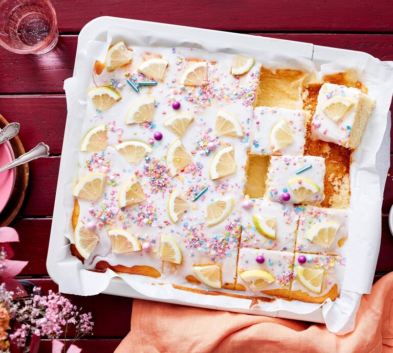 Auf einem weißen Teller liegt ein saftiger Zitronen-Buttermilchkuchen. Er ist mit Puderzuckerglasur und Zuckerstreuseln dekoriert und mit frischen Zitronen bedeckt.  | © EAT CLUB/Isabell Triemer