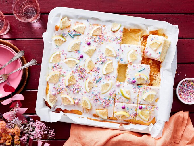 Auf einem weißen Teller liegt ein saftiger Zitronen-Buttermilchkuchen. Er ist mit Puderzuckerglasur und Zuckerstreuseln dekoriert und mit frischen Zitronen bedeckt.  | © EAT CLUB/Isabell Triemer