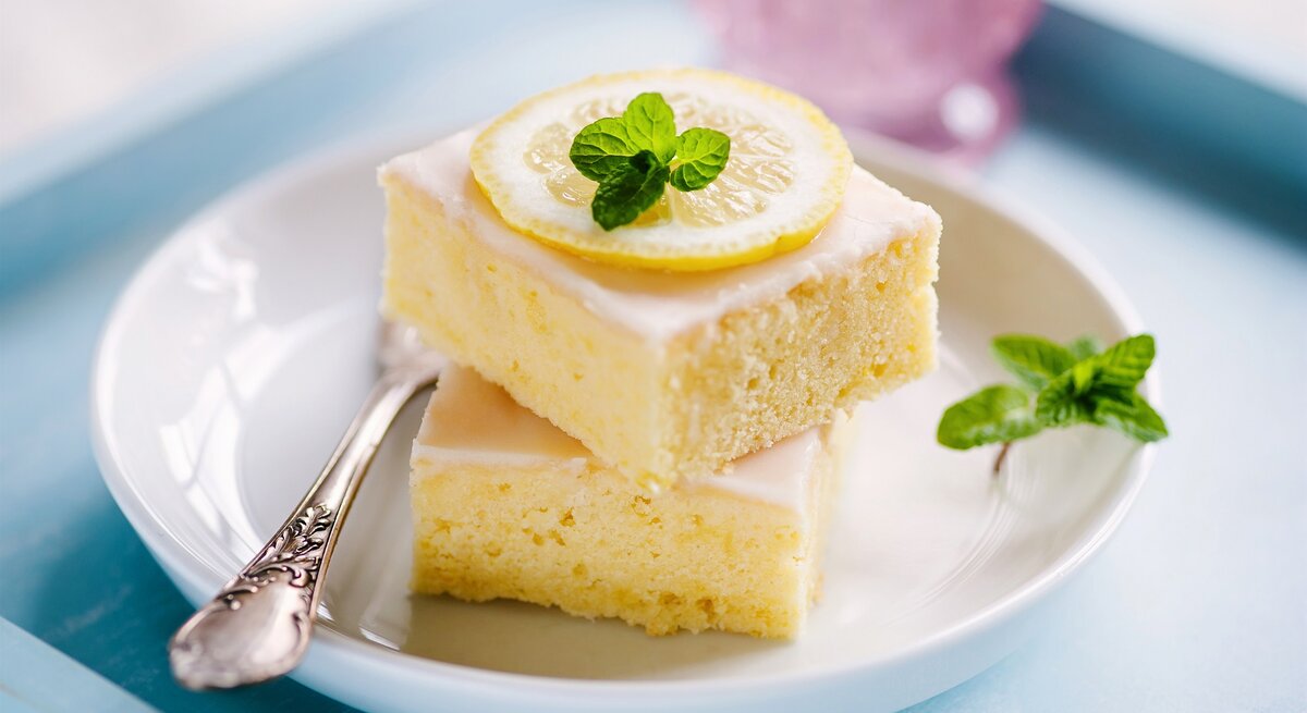 Zwei quadratische Stücke Zitronenkuchen auf einem weißen Teller. 