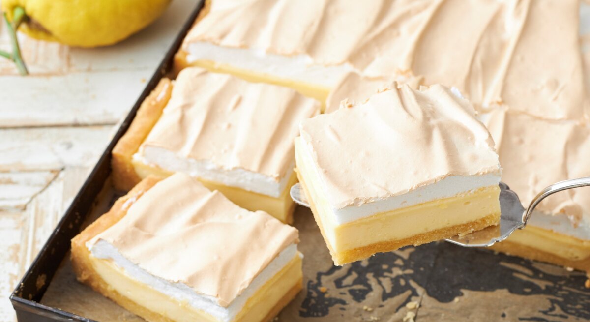 Zitronen-Schmand-Kuchen mit Baiser in einem dunklen Blech auf einem hellen Tisch mit frischen Zitronen. 