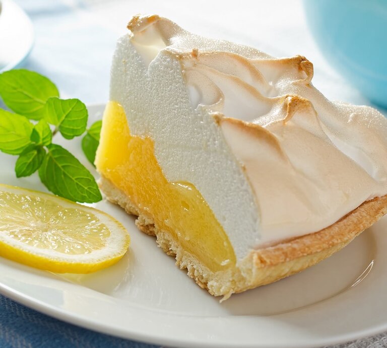 Ein Stück von dem Zitronenkuchen auf einem weißen Teller und einer weißblauen Tischdecke. Auf dem Teller ist auch ein Minzzweig und eine Zitronenscheibe. | © Shutterstock/ Foodio