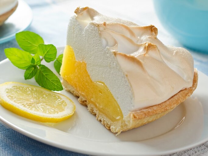Ein Stück von dem Zitronenkuchen auf einem weißen Teller und einer weißblauen Tischdecke. Auf dem Teller ist auch ein Minzzweig und eine Zitronenscheibe. | © Shutterstock/ Foodio
