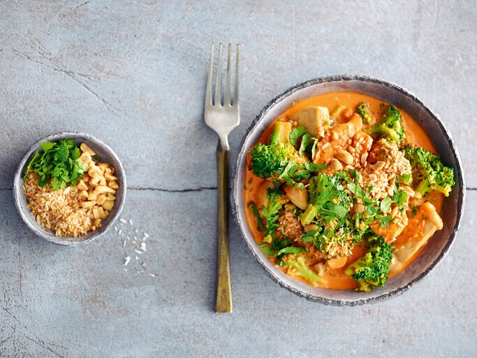Ein Teller Hähnchen-Brokkoli-Curry in der Draufsicht, daneben eine Gabel und eine Schale mit Erdnüssen und Kräutern. | © StockFood/ Becker Joest Volk Verlag/ Hubertus Schüler