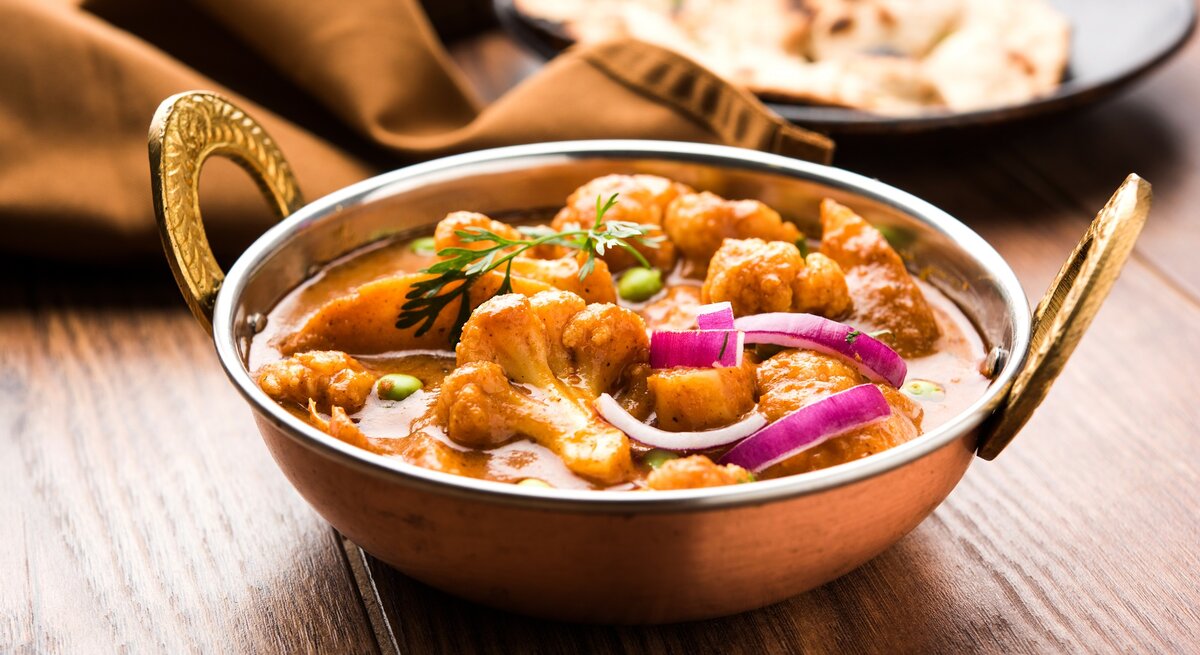 Eine Schüssel Aloo Gobi, ein Kartoffel-Blumenkohl-Curry, mit roter Zwiebel und Kräutern, im Hintergrund ein Naan-Brot.