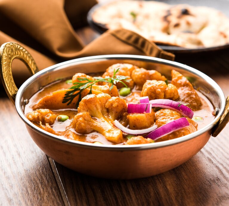 Eine Schüssel Aloo Gobi, ein Kartoffel-Blumenkohl-Curry, mit roter Zwiebel und Kräutern, im Hintergrund ein Naan-Brot. | © Adobe Stock/ StockImageFactory