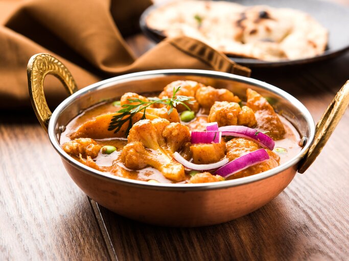 Eine Schüssel Aloo Gobi, ein Kartoffel-Blumenkohl-Curry, mit roter Zwiebel und Kräutern, im Hintergrund ein Naan-Brot. | © Adobe Stock/ StockImageFactory