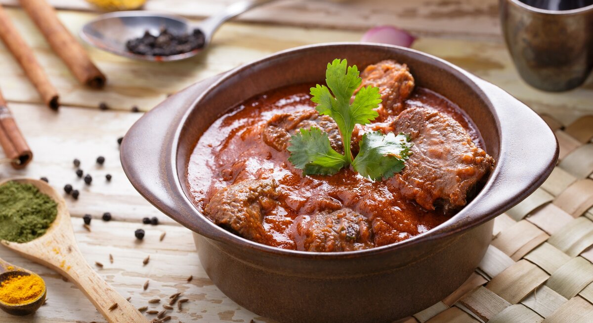 Draufsicht, schräg: Ein Tontopf mit dem asiatischen Currygericht Rogan Josh steht auf einer Bastmatte, die auf einem Holztisch liegt. Drumherum liegen Holzlöffel mit Gewürzen.