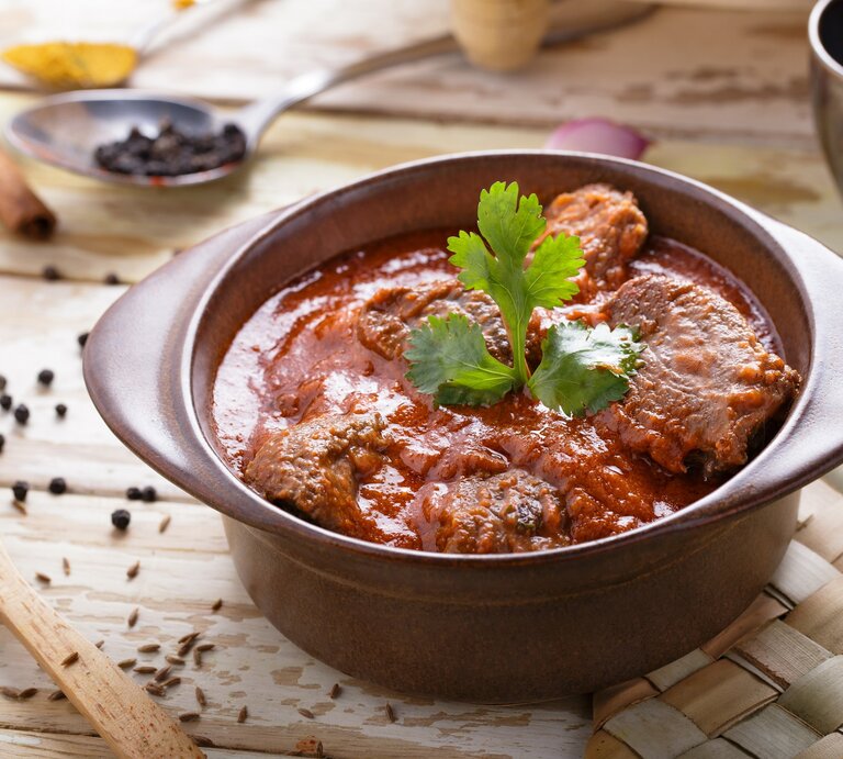 Draufsicht, schräg: Ein Tontopf mit dem asiatischen Currygericht Rogan Josh steht auf einer Bastmatte, die auf einem Holztisch liegt. Drumherum liegen Holzlöffel mit Gewürzen. | © Shutterstock / Odua Images