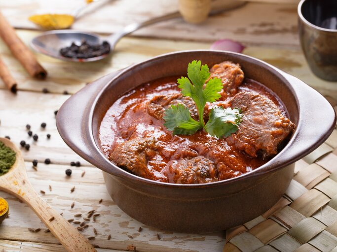 Draufsicht, schräg: Ein Tontopf mit dem asiatischen Currygericht Rogan Josh steht auf einer Bastmatte, die auf einem Holztisch liegt. Drumherum liegen Holzlöffel mit Gewürzen. | © Shutterstock / Odua Images