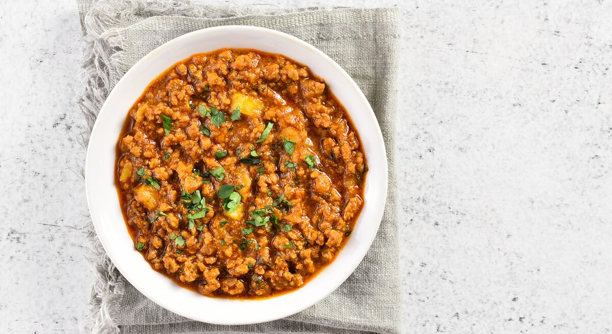 Eine weiße Schale mit dem rötlichen Curry auf einem hell-beigenen Leinentuch und einem weißen Tisch.