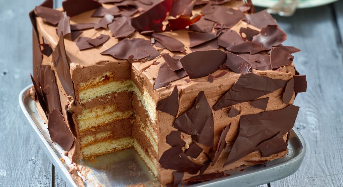 Angeschnittene viereckige Schoko-Sahne-Torte mit Schoko-Scherben verziert auf Metall-Präsentierteller, heller Hintergrund unscharf. 
