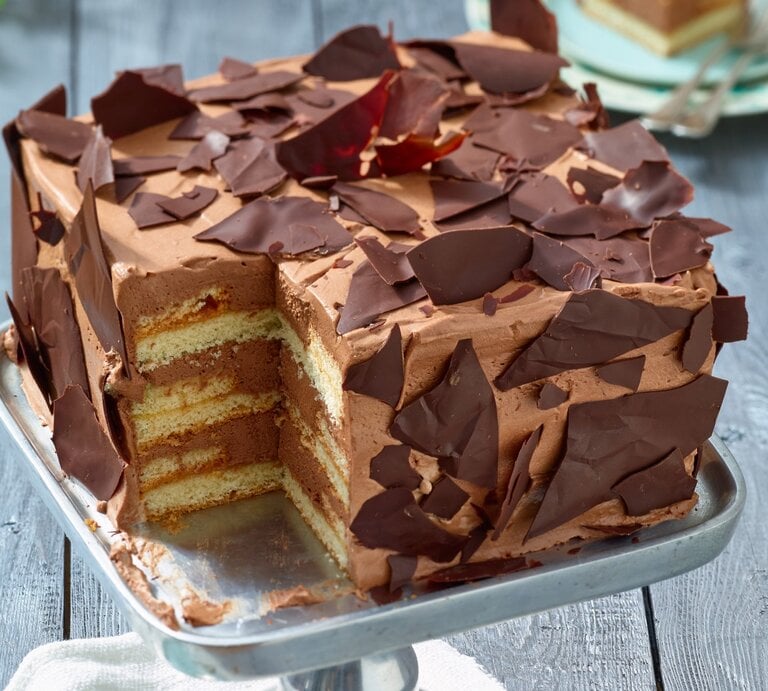 Angeschnittene viereckige Schoko-Sahne-Torte mit Schoko-Scherben verziert auf Metall-Präsentierteller, heller Hintergrund unscharf.  | © EAT CLUB / Isabell Triemer