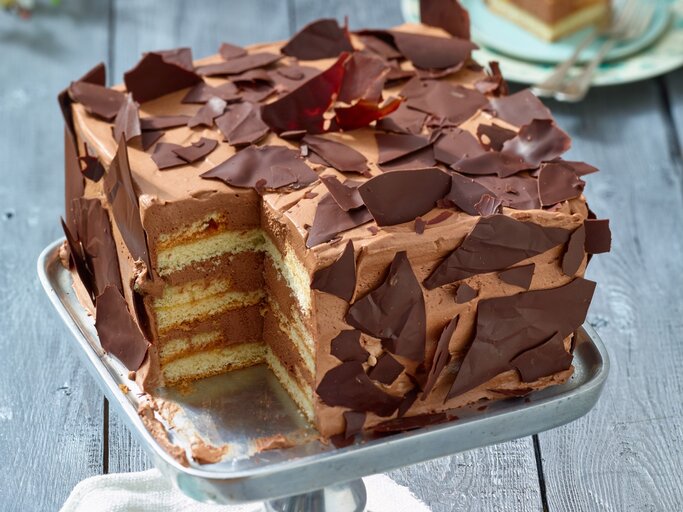 Angeschnittene viereckige Schoko-Sahne-Torte mit Schoko-Scherben verziert auf Metall-Präsentierteller, heller Hintergrund unscharf.  | © EAT CLUB / Isabell Triemer