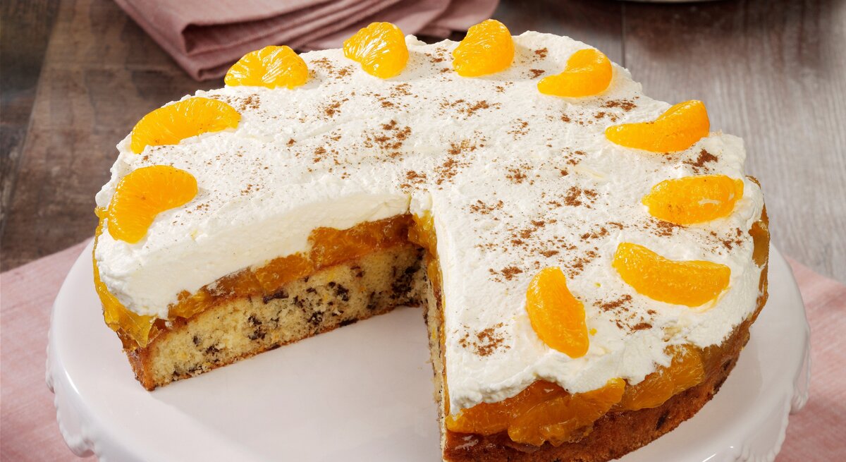 Eine Mandarinen-Sahnetorte mit Schokoraspeln und Schmand auf einem Kuchentablett. Die Torte ist bereits angeschnitten und zwei Stücke wurden heraus genommen.