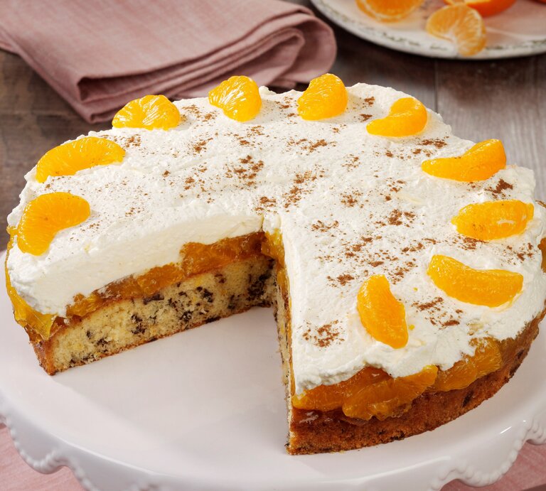 Eine Mandarinen-Sahnetorte mit Schokoraspeln und Schmand auf einem Kuchentablett. Die Torte ist bereits angeschnitten und zwei Stücke wurden heraus genommen. | © StockFood Studios