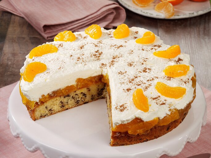 Eine Mandarinen-Sahnetorte mit Schokoraspeln und Schmand auf einem Kuchentablett. Die Torte ist bereits angeschnitten und zwei Stücke wurden heraus genommen. | © StockFood Studios