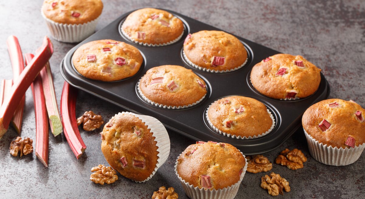 Rhabarber-Muffins mit Walnüssen in einer Muffinform, einige Muffins liegen daneben, außerdem frische Rhabarberstangen.
