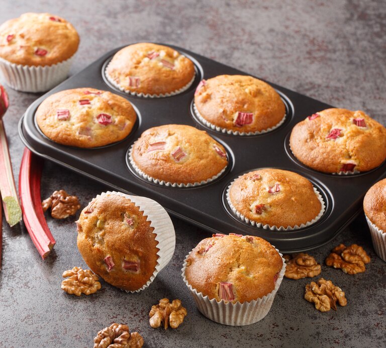 Rhabarber-Muffins mit Walnüssen in einer Muffinform, einige Muffins liegen daneben, außerdem frische Rhabarberstangen. | © Getty Images/ ALLEKO