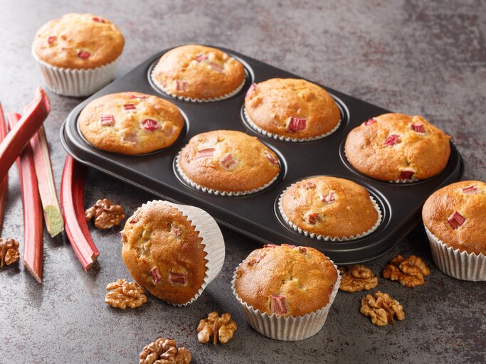 Rhabarber-Muffins mit Walnüssen in einer Muffinform, einige Muffins liegen daneben, außerdem frische Rhabarberstangen. | © Getty Images/ ALLEKO