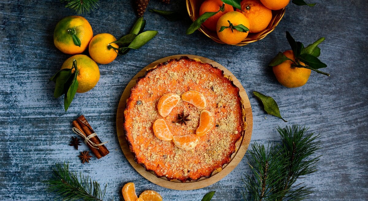 Ein Kürbiskuchen mit Mandarinen ist mit gehackten Pistazien und Mandarinen angerichtet. Der Kuchen steht auf einem blaugrauen Untergrund. Um den Kuchen herum liegen Zimt, Mandarinen ind Tannenäste als Dekoration.