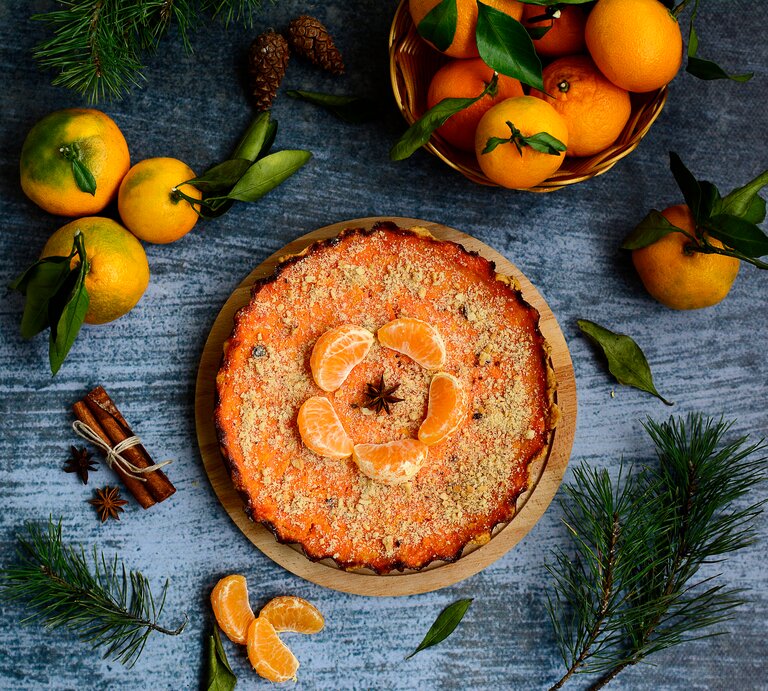 Ein Kürbiskuchen mit Mandarinen ist mit gehackten Pistazien und Mandarinen angerichtet. Der Kuchen steht auf einem blaugrauen Untergrund. Um den Kuchen herum liegen Zimt, Mandarinen ind Tannenäste als Dekoration. | © Shutterstock / Valeriya Zeleneva