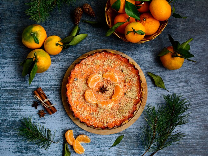 Ein Kürbiskuchen mit Mandarinen ist mit gehackten Pistazien und Mandarinen angerichtet. Der Kuchen steht auf einem blaugrauen Untergrund. Um den Kuchen herum liegen Zimt, Mandarinen ind Tannenäste als Dekoration. | © Shutterstock / Valeriya Zeleneva