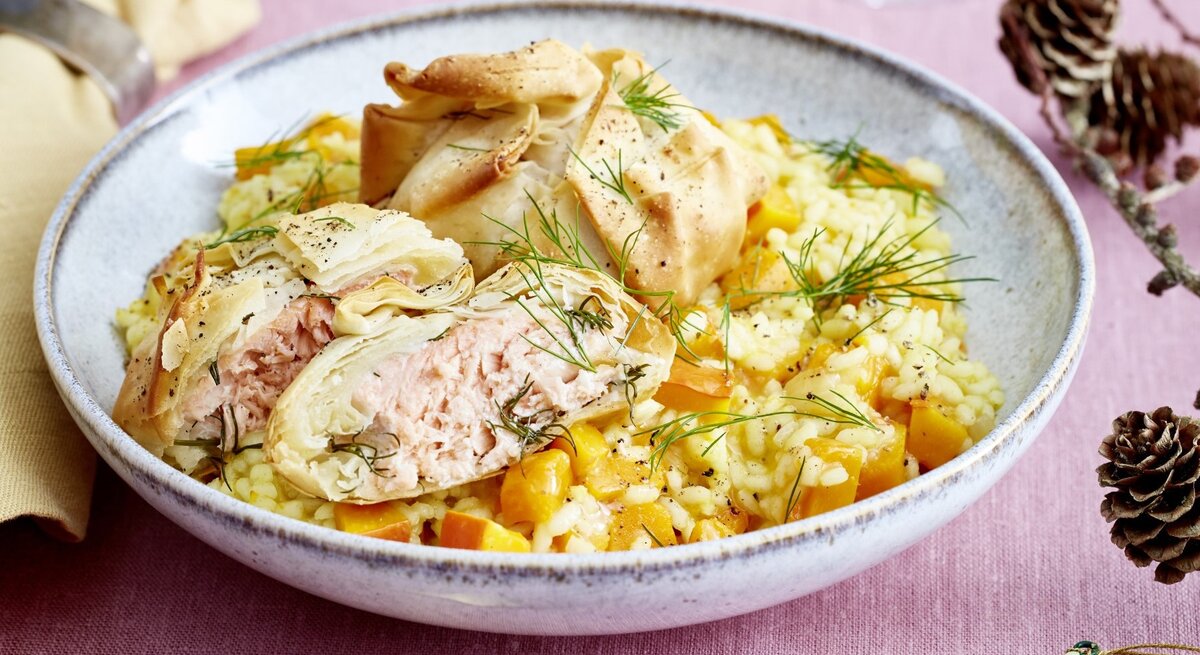 Lachs-Päckchen auf Kürbis-Risotto auf grauem Teller und rosa Tischdecke. Daneben Serviette und Dekoration. Draufsicht.