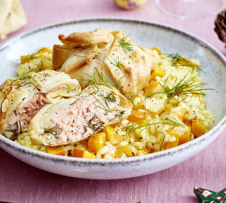 Lachs-Päckchen auf Kürbis-Risotto auf grauem Teller und rosa Tischdecke. Daneben Serviette und Dekoration. Draufsicht. | © EAT CLUB / Isabell Triemer