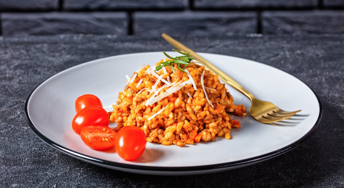 Hackfleisch-Risotto mit Cocktailtomaten auf weißem Teller mit Kupfergabel auf dunklem Untergrund. Draufsicht.