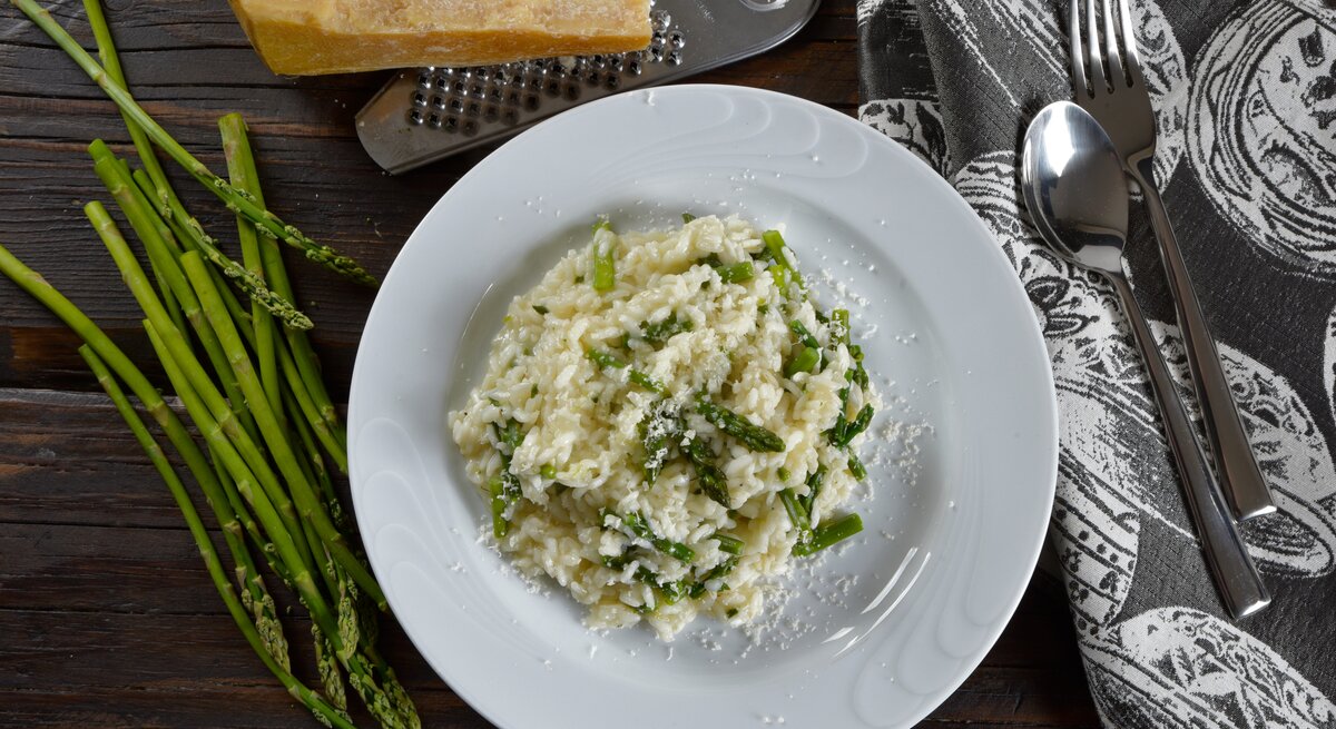 Draufsicht: Eine weißer Teller Wildspargel-Risotto steht auf einem dunklen Untergrund. Drum herum liegen wilder Spargel, Parmesan, ein Reibe, Besteck und ein Tuch.