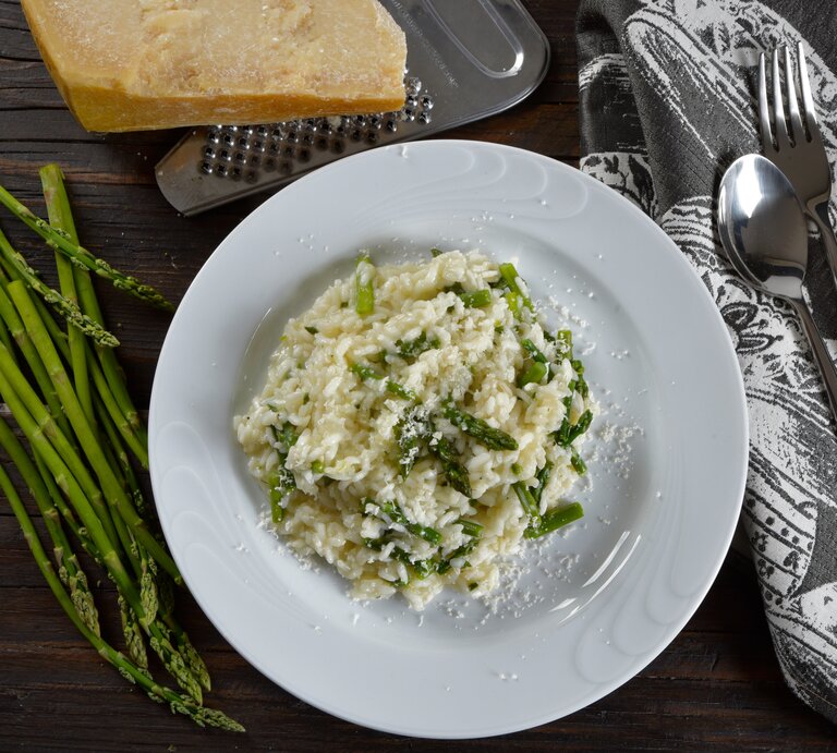 Draufsicht: Eine weißer Teller Wildspargel-Risotto steht auf einem dunklen Untergrund. Drum herum liegen wilder Spargel, Parmesan, ein Reibe, Besteck und ein Tuch. | © Shutterstock / barbajones