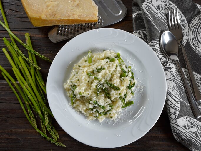 Draufsicht: Eine weißer Teller Wildspargel-Risotto steht auf einem dunklen Untergrund. Drum herum liegen wilder Spargel, Parmesan, ein Reibe, Besteck und ein Tuch. | © Shutterstock / barbajones