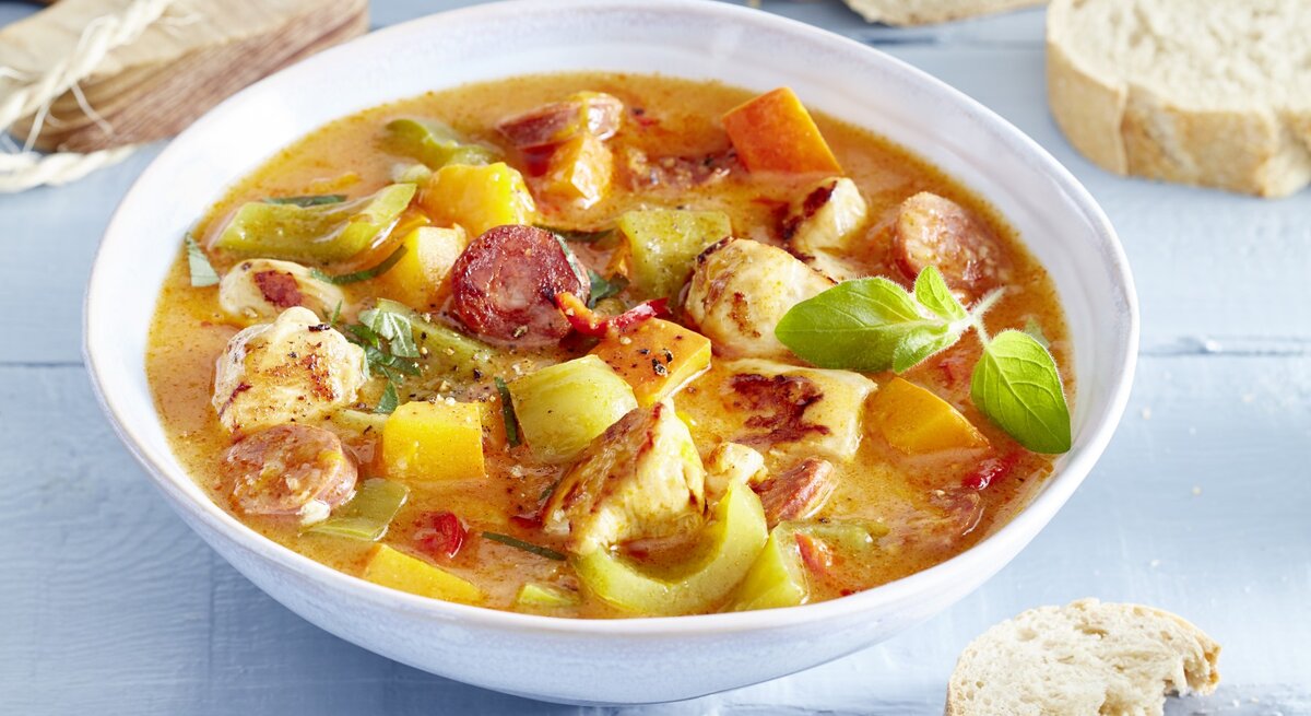 Hähnchen-Kürbis-Gulasch in einem weißten tiefen Teller. Daneben liegt Brot und ein Holzbrett. 