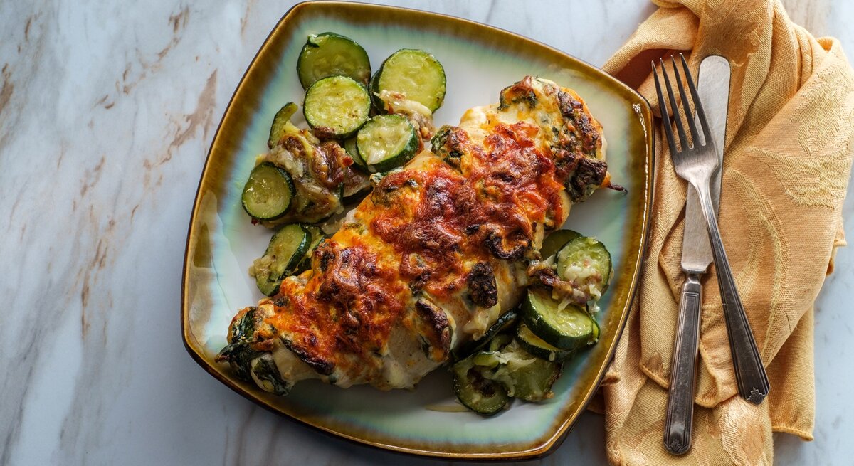 Hasselback Hähnchen mit Zucchini und Spinat auf einem farbigen Teller. Daneben Besteck auf einem Tuch. 