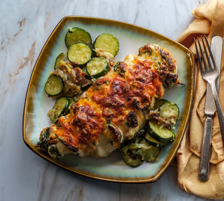 Hasselback Hähnchen mit Zucchini und Spinat auf einem farbigen Teller. Daneben Besteck auf einem Tuch.  | © GettyImages/EzumeImages