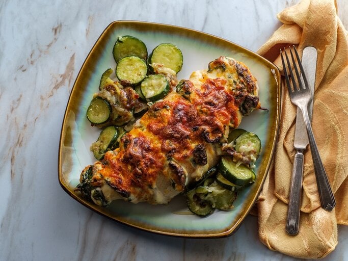 Hasselback Hähnchen mit Zucchini und Spinat auf einem farbigen Teller. Daneben Besteck auf einem Tuch.  | © GettyImages/EzumeImages