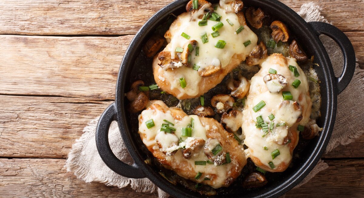Überbackene Hähnchenbrust mit Pilzen, Lauch und Mozzarella in schwarzer Gussform mit Henkeln auf Leinentuch und Holztisch. Aufnahme aus der Vogelperspektive.  