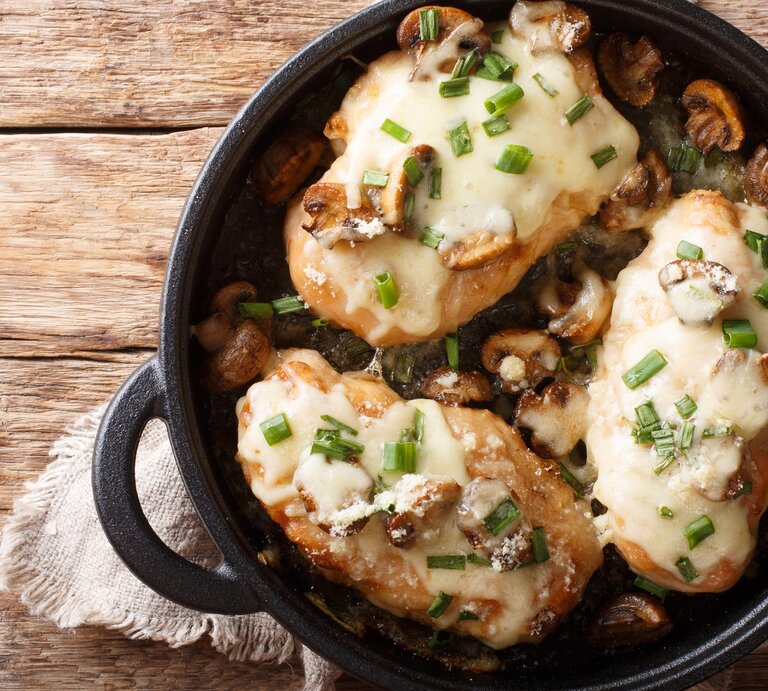 Überbackene Hähnchenbrust mit Pilzen, Lauch und Mozzarella in schwarzer Gussform mit Henkeln auf Leinentuch und Holztisch. Aufnahme aus der Vogelperspektive.   | © Getty Images/ALLEKO
