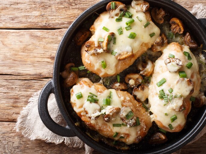 Überbackene Hähnchenbrust mit Pilzen, Lauch und Mozzarella in schwarzer Gussform mit Henkeln auf Leinentuch und Holztisch. Aufnahme aus der Vogelperspektive.   | © Getty Images/ALLEKO