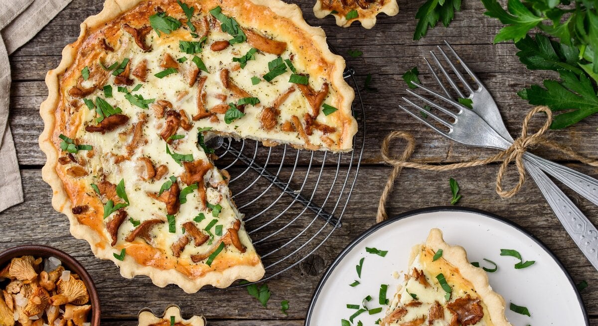 Eine angeschnittene Pfifferling-Quiche mit Schmand und Bergkäse steht auf einem Kuchengitter. Daneben liegen Besteck sowie ein kleiner Teller mit einem Stück Quiche.