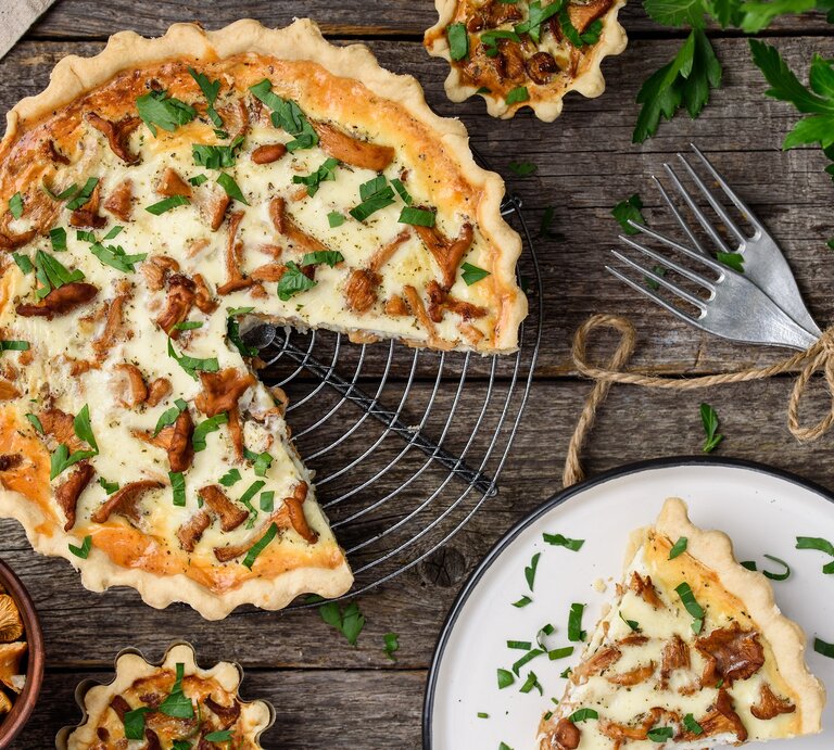 Eine angeschnittene Pfifferling-Quiche mit Schmand und Bergkäse steht auf einem Kuchengitter. Daneben liegen Besteck sowie ein kleiner Teller mit einem Stück Quiche. | © Shutterstock / rom_olik