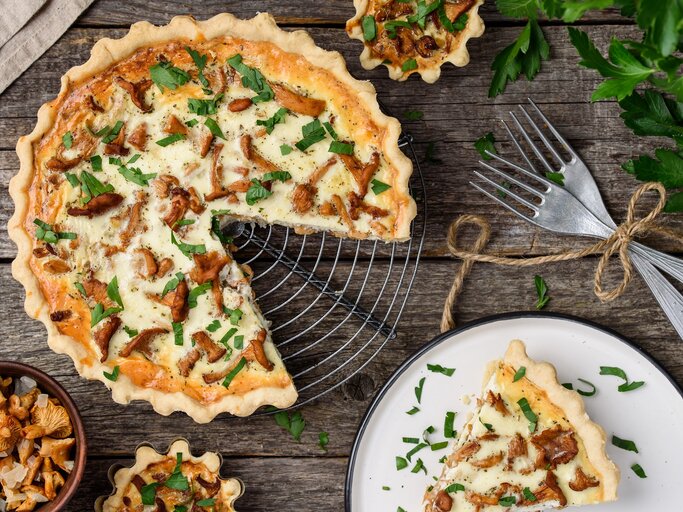 Eine angeschnittene Pfifferling-Quiche mit Schmand und Bergkäse steht auf einem Kuchengitter. Daneben liegen Besteck sowie ein kleiner Teller mit einem Stück Quiche. | © Shutterstock / rom_olik