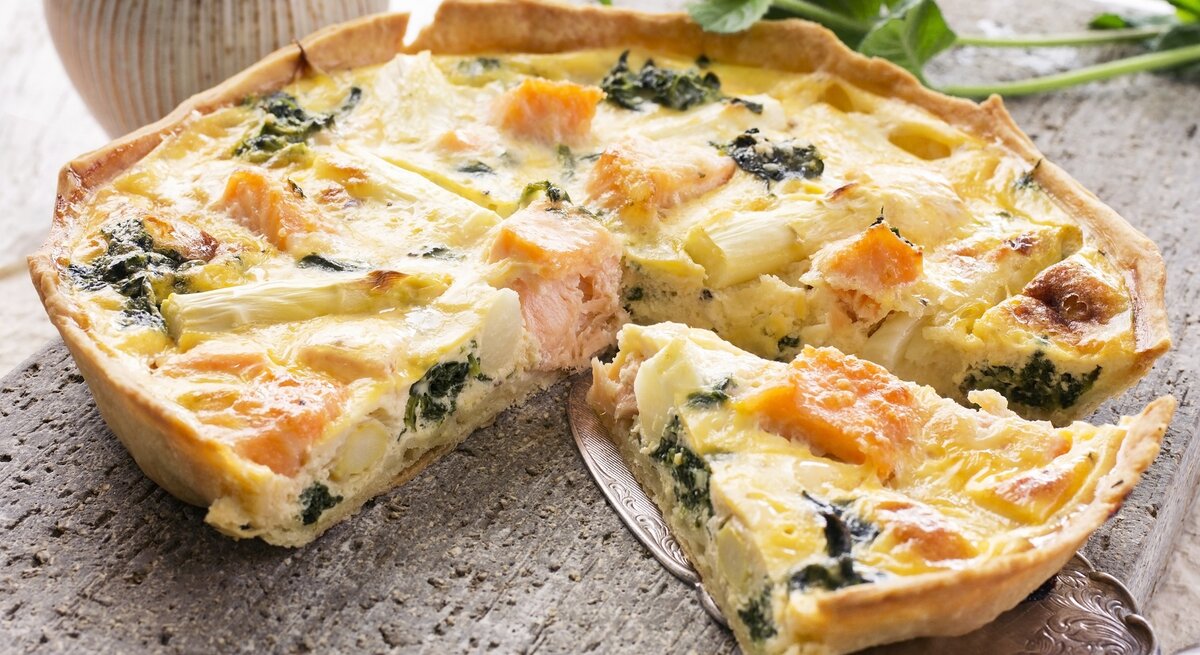 Die portionierte Quiche auf einem Steintisch mit einem Tortenheber, der ein Stück etwas anhebt. Im Hintergrund Vasen und Pflanzen.