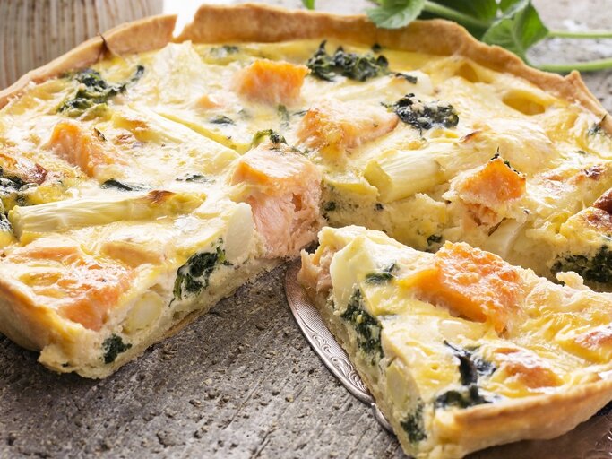 Die portionierte Quiche auf einem Steintisch mit einem Tortenheber, der ein Stück etwas anhebt. Im Hintergrund Vasen und Pflanzen. | © Shutterstock/ hlphoto