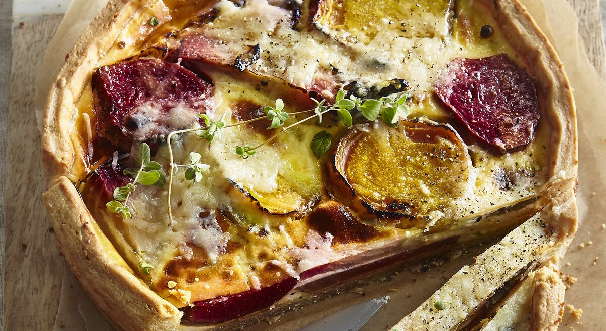 Angeschnittene Quiche mit Thymianzweig, geschmolzenem Käse auf Backpapier und geteiltem Holzbrett mit Steingutrand aus der Vogelperspektive. Rote Bete und Gelbe Bete in Scheiben sichtbar. Der Rand der Quiche leicht erhöht und gebräunt. Zwei Scheiben längs aufgeschnitten. Messer mit silberner Klinge und Holzgriff zwischen den Scheiben aufgelegt.
